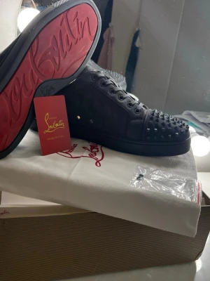 Christian Louboutin svarta sneakers - Svarta sneakers från Christian Louboutin med klassisk röd sula och nitar på tån. Skorna har lågt skaft, Gråa skosnören och är tillverkade i mocka. Perfekta för dig som vill sticka ut med en lyxig och edgy stil. Skorna har använts ett tag och 4-7 spikar är borta men man får med en ny spik. De är i okej skicka, därför de säljs billigt. Box och allt medföljer. Passar inte priset är det bara att hojta till. 