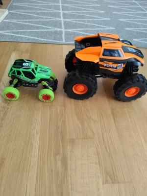 Två leksaksbilar monstertruckar - Säljer två coola leksaksbilar i monstertruck-stil. En är grön med svarta detaljer och röda hjul, och den andra är orange och svart med stora hjul och texten 'Power 05' på sidan. Perfekta för lek på golvet och för dig som gillar fartfyllda bilar.