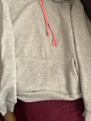Grå hoodie med rosa detaljer - Grå hoodie med rosa snören och en stor magficka. På huvan finns ett coolt rosa tryck i graffiti-stil. Hoodien har ribbade muddar och är perfekt för en avslappnad stil.