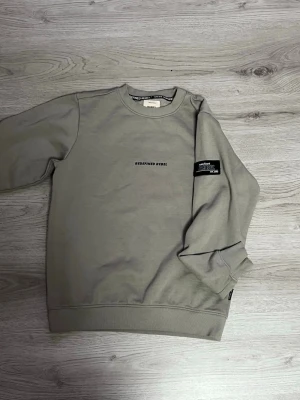Grå sweatshirt från Redefined Rebel - Snygg grå sweatshirt från Redefined Rebel med tryckt logga på bröstet och en svart patch på ärmen. Tröjan har rund halsringning och ribbade muddar vid ärmslut och nederkant. Perfekt för en avslappnad och trendig stil.
