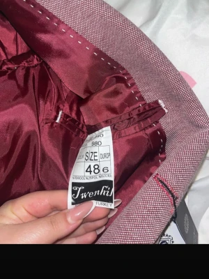Vinröd kavaj från Twenhi, storlek 48 - Snygg vinröd kavaj från Twenhi i storlek 48. Kavajen har ett diskret mönster, två knappar framtill och innerfoder i matchande vinröd nyans. Materialet är 70% ull, 25% polyester och 5% lycra för en bekväm och stilren look.
