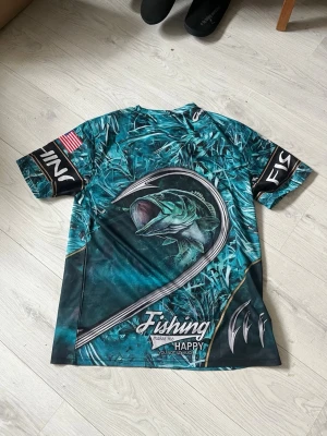 Fisketee med fiskemotiv och tryck - Cool t-shirt från FINS med fiskemotiv och texten 'Fishing makes me happy'. T-shirten är blågrön med tryck av fisk och krokar, samt detaljer på ärmarna. Perfekt för dig som gillar fiske och vill sticka ut med en unik stil.