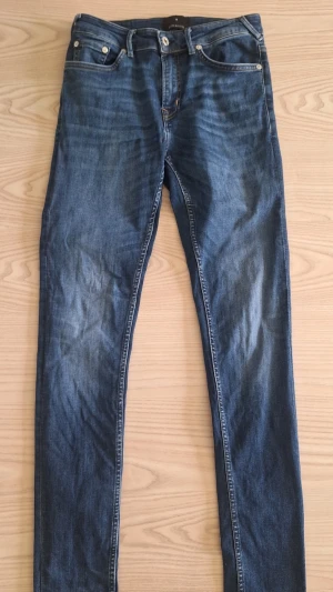 Blå skinny fit jeans från JUNK de LUXE W30 L32 - Snygga blå skinny fit jeans från JUNK de LUXE, J-01, med klassisk femficksdesign och diskreta slitningar. Jeansen har normal midja och stängs med dragkedja och knapp. Perfekta för en avslappnad och trendig look.