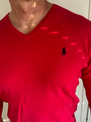 Röd V-ringad tröja från Ralph Lauren - Snygg röd tröja från Ralph Lauren med V-ringning och klassisk broderad logga på bröstet. Långärmad modell i mjukt material, perfekt för en stilren look. Passar till både jeans och chinos.