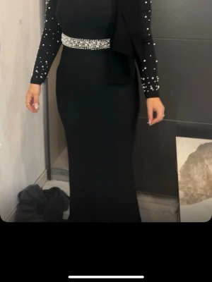 Svart långklänning med pärldetaljer - Elegant svart långklänning med figurnära passform och pärldetaljer vid midjan och på ärmarna. Klänningen har långa ärmar och en klassisk, stilren look som passar perfekt till speciella tillfällen.