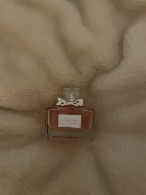 Miss Dior Absolutely Blooming parfym - Miss Dior Absolutely Blooming parfym med en elegant glasflaska och en söt silverfärgad rosett vid halsen. Parfymen har en rosa nyans och flaskan har en stilren, fyrkantig form med genomskinligt lock. Perfekt för dig som gillar lyxiga dofter.