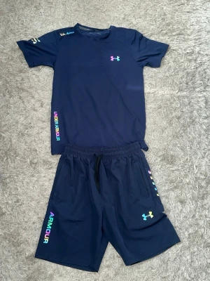 Under Armour marinblått (reflex) - Snyggt marinblått träningsset från Under Armour med t-shirt och shorts. Setet har färgglada loggor och textdetaljer i regnbågsfärger. Tillverkat i ett lätt och snabbtorkande material som passar perfekt för träning och sportaktiviteter. Det står att det är storlek M men passformen är för storlek S