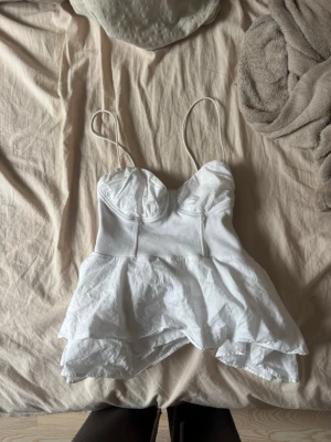 Vit volang topp från Zara - Supersöt vit topp från Zara med tunna axelband och volang nedtill. Toppen har markerade sömmar och bystparti som ger en korsettkänsla. Perfekt för sommarens alla tillfällen och lätt att styla med jeans eller kjol.