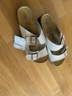Beige Birkenstock sandaler med spännen - Snygga beige sandaler från Birkenstock med två breda remmar i mocka och justerbara metallspännen. Fotbädden är mjuk och ergonomisk för extra komfort. Perfekta för en avslappnad och trendig stil.