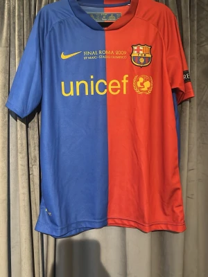 FC Barcelona Final Roma 2009 tröja - Säljer en officiell FC Barcelona matchtröja från Final Roma 2009, med klassisk halvblå och halvröd design. Tröjan har klubbmärke, Nike-logga och Unicef-tryck i gult framtill. Tillverkad i lätt och ventilerande polyester, perfekt för fotboll eller samling.