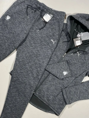 Grå tracksuit Puma - Mörkgrå melerade mjukisbyxor och luvtröja från Puma med dragkedja och broderad logga. Båda delarna har en regular fit-modell och är tillverkade i ett mjukt och stretchigt material (Puma DRYCELL) som passar perfekt för chill eller träning till ett galet pris.