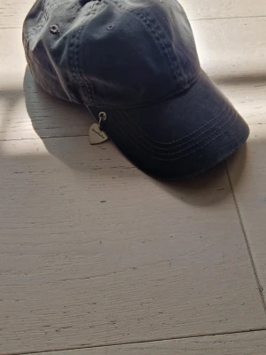Svart keps från Acne Studios - Snygg svart keps från Acne Studios med böjd skärm och coolt hjärtformat metallhänge på sidan där märkesnamnet står. Kepsen är i bomull och har klassisk sexpanelsdesign med synliga sömmar. Perfekt accessoar för en trendig look.