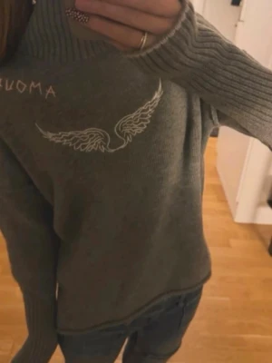 Grå stickad tröja med vingtryck - Mysig grå stickad tröja med höga ribbade muddar och ett vitt vingtryck på bröstet. Tröjan har även textdetalj vid axeln och en hög krage. Perfekt för dig som gillar unika detaljer och bekväma plagg.