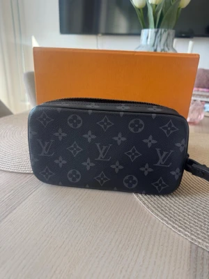 Louis Vuitton svart monogram handväska - Snygg svart handväska från Louis Vuitton med klassiskt monogrammönster i grått. Väskan har en rektangulär form, dragkedja upptill och en svart kedjedetalj framtill. Tillverkad i canvas med läderdetaljer. Perfekt för dig som gillar lyxiga accessoarer.