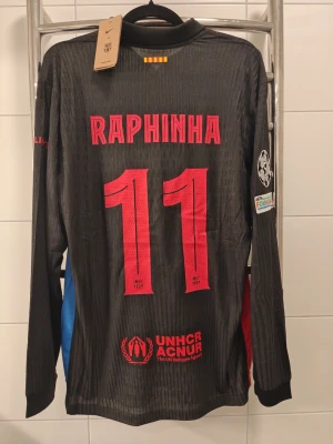 FC Barcelona långärmad spelar Version matchtröja Raphinha - Snygg svart långärmad spelar Version FC Barcelona matchtröja från Nike med Raphinha och nummer 11 på ryggen. Tröjan har röda och blå detaljer på ärmarna, klubbmärke och sponsorloggor på bröstet samt Champions League-märke på ärmen. Perfekt för fotbollsfans!