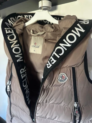 Beige Väst Moncler - Stilren beige dunväst från Moncler med svart dragkedja och stora logotypband längs huvan. Västen har två framfickor med dragkedja och Moncler-logga på bröstet. Perfekt för lager-på-lager och streetstyle. Storlek XL men skulle säga att den passar M.