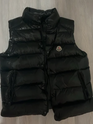 Svart dunväst från Moncler - Snygg svart dunväst från Moncler med hög krage och två dragkedjefickor framtill. Västen har ett quiltat mönster och Moncler-logga på bröstet. Perfekt för lager på lager och håller dig varm med sin fluffiga dunfyllning.
