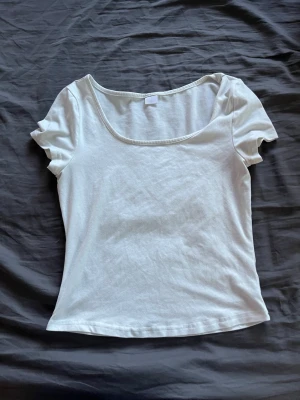 Vit basic topp med rund hals - En enkel och stilren vit topp med rundad, djup urringning och korta ärmar. Tillverkad i mjuk bomull som känns skön mot huden. Perfekt att matcha med jeans eller kjol för en clean look.