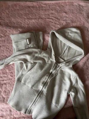 Grått loungewear-set med huva - Mysigt loungewear-set i ljusgrått med ziptröja och matchande byxor. Tröjan har huva, dragkedja och bred ribbad mudd vid ärmslut och midja. Byxorna har ribbade muddar och dekorativt spänne. Perfekt för avslappnade dagar hemma.