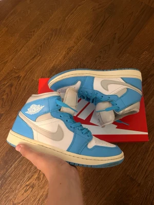 Nike Air Jordan 1 midblå/vit - Säljer ett par Nike Air Jordan 1 High sneakers i blått och vitt. Skorna har klassisk hög siluett, vita läderpaneler och ljusblå overlays samt swoosh i grått. Ikonisk Air Jordan-logga på sidan och matchande blå skosnören. Perfekta för dig som gillar streetwear.