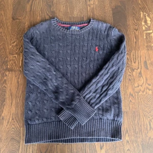 Mörkblå kabelstickad tröja Polo - Mörkblå kabelstickad tröja från Polo Ralph Lauren med rund halsringning och röd broderad logga på bröstet. Tröjan har ribbstickade muddar vid ärmslut och nederkant, perfekt för en klassisk och stilren look.