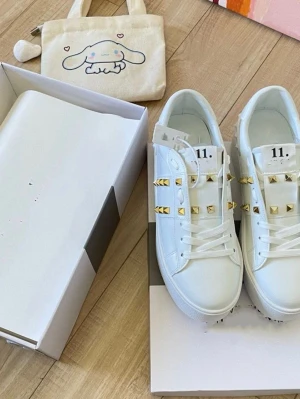 Valentino Rockstud Untitled vita sneakers - Snygga vita sneakers från Valentino med ikoniska guldiga nitar på sidorna och ovandelen. Skorna har en klassisk låg siluett, vita skosnören och detaljer i läder. Perfekta för dig som vill sticka ut med en lyxig touch.