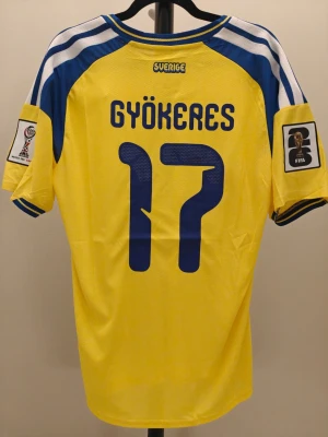 Spelar version Sverige matchtröja Gyökeres #17 Adidas - Gul och blå Sverige matchtröja i spelar Version med Gyökeres och nummer 17 på ryggen. Tröjan har FIFA och VM-märken på ärmarna, broderad svensk landslagssköld och ventilerande meshmaterial. Perfekt för fotbollsfans som vill stötta landslaget.