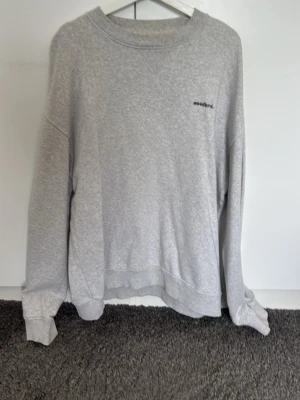 Grå sweatshirt från Woodbird - Snygg grå sweatshirt från Woodbird med minimalistisk svart logga på bröstet. Tröjan har rund halsringning, långa ärmar och är gjord i mjukt material som känns skönt mot huden. Perfekt för en avslappnad och stilren look.