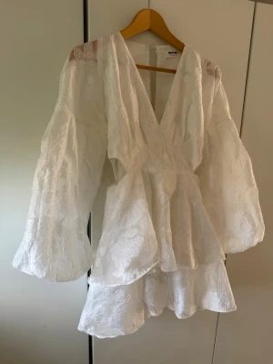 Studentklänning vit klänning - Superfin vit kort omlottklänning från showpo med volanger och långa puffärmar. Klänningen har v-ringning och är tillverkad i ett lätt strukturerat tyg med subtilt blommönster. Perfekt för sommarens alla tillfällen! Ihop sydd vid brösten för var öppen hela vägen