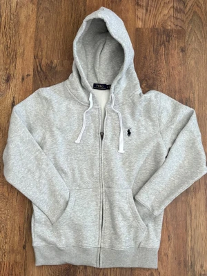 Grå hoodie från Polo Ralph Lauren - Snygg ljusgrå hoodie från Polo Ralph Lauren med dragkedja, huva och vita dragsnören. Klassisk logga broderad på bröstet och kängurufickor framtill. Tillverkad i mjuk bomullsblandning som är perfekt för chill dagar.