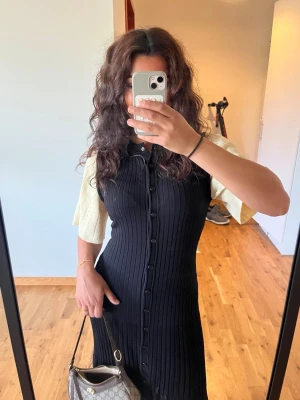 Sandro klänning  - Svart ribbad klänning med knappar framtill och kort ärm. Klänningen har en figurnära passform och är knälång. Perfekt för dig som gillar en stilren och enkel look.