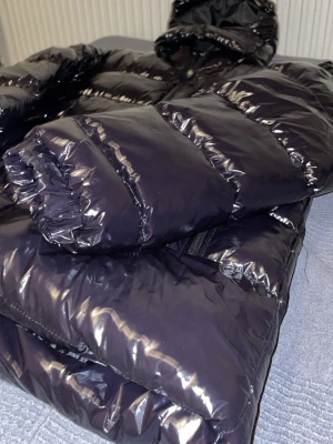 Svart pufferjacka Polo Ralph Lauren - Snygg svart pufferjacka från Polo Ralph Lauren i glansigt material. Jackan har huva och breda kanaler som ger en riktigt puffig look. Perfekt för kalla vinterdagar och håller dig varm med stil.