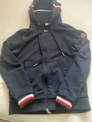 Moncler zip - Snygg mörkblå hoodie från Moncler med dragkedja, huva och dragsko. Röd, vit och blå randdetalj på huva och ärmslut. Klassisk Moncler-logga på ena ärmen. Perfekt för en chill och sportig stil. lite solbränd vid luvan