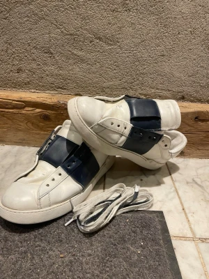 Valentino Open sneakers med blå detalj - Snygga vita sneakers från Valentino med bred mörkblå rem över vristen istället för klassiska snören. Skorna är i läder och har en chunky sula samt nitar bak på hälen för en cool touch. Perfekta för dig som gillar stilrena men ändå unika sneakers. En del slitna.