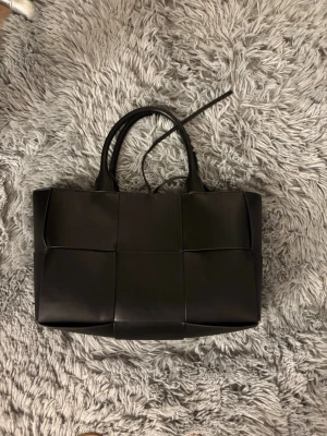 Svart flätad toteväska i läderstil - Stilren svart handväska i rektangulär tote-modell med breda flätade paneler. Väskan har två rejäla handtag och är tillverkad i ett material som liknar läder. Perfekt för dig som gillar minimalistisk och trendig design. Använt nån gång, fortfarande i bra skick kan vara dammig 