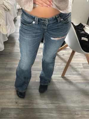 Blå bootcut jeans med slitning - Säljer ett par blå bootcut jeans med slitning på låret och snygga broderade detaljer på bakfickan. Jeansen har klassisk femficksmodell och är tillverkade i denim med lätt stretch. Perfekta för en avslappnad och trendig look. Från Gina 14+. Lite slitningar där nere men inget man tänker på 