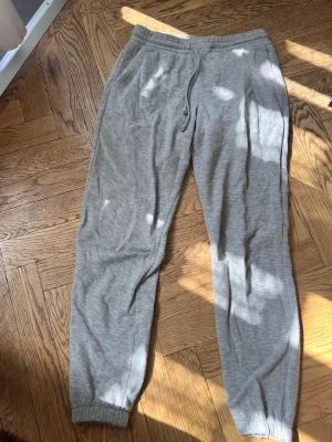 Grå mjukisbyxor med dragsko - Ett par klassiska grå mjukisbyxor med elastisk midja och dragsko. Byxorna har ribbade muddar vid bensluten och sidofickor. Perfekta för chill eller träning, tillverkade i mjukt material som känns skönt mot huden.