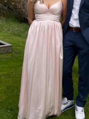 Ljusrosa långklänning med snörning - Elegant ljusrosa långklänning med v-ringad överdel och smala axelband. Klänningen har snörning i ryggen och är tillverkad i ett glansigt material som ger en lyxig känsla. Perfekt för speciella tillfällen.