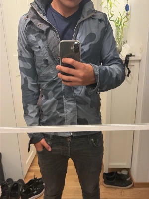 Blå camo vindjacka med huva - Snygg vindjacka i blått camomönster med dragkedja och tryckknappar. Jackan har huva, snedställda fickor med dragkedja och justerbara ärmslut. Perfekt för blåsiga dagar och streetwear-stil.