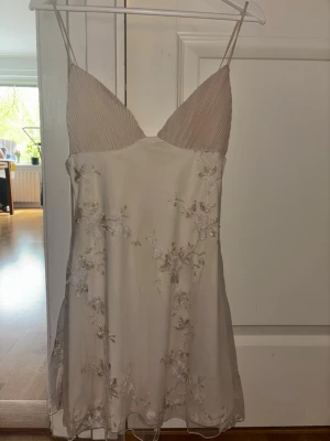 Krämvit blommig studentklänning - Söt kort klänning i krämvit satin med tunna axelband och v-ringad byst. Klänningen har broderade blommor i vitt och guld, samt lätt utsvängd kjol. Köpt på Vinted men säljer den då den var lite för liten för mig💕