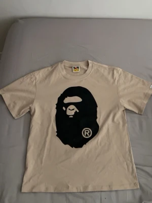 Beige BAPE t-shirt med ape-print - Snygg beige t-shirt från BAPE med det ikoniska svarta ape-huvudet tryckt på bröstet. Klassisk passform och rund halsringning. Perfekt för dig som gillar streetwear och vill sticka ut med en stilren look.