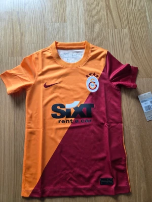 Nike Galatasaray hemmatröja 2021/22 unisex barn storlek XS , 122-128 cm - Fotbollströja från Galatasaray i orange och vinrött. Tröjan har klubbens emblem på bröstet tillsammans med fyra stjärnor. På framsidan finns texten "SIXT rent a car" och på ryggen "MAGDEBURGER SIGORTA". Nike-logotypen finns på höger sida av bröstet.