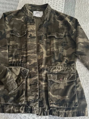 Camo skepparkavaj från ONLY L - Snygg skepparkavaj i camouflage-mönster från ONLY. Jackan har fyra fickor framtill, ståkrage och knäppning med knappar. Tillverkad i ett slitstarkt bomullstyg, perfekt för en cool streetstil.