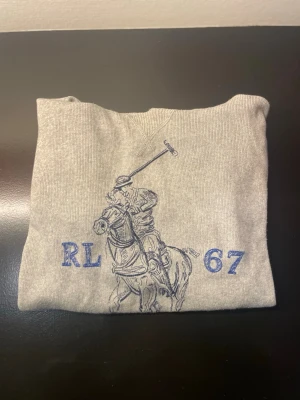 Grå hoodie från Ralph Lauren - Snygg grå hoodie från Ralph Lauren med blått broderi framtill som visar en polospelare och texten RL 67. Klassisk huva och mjukt material som känns skönt mot huden. Perfekt för dig som gillar stilrena och sportiga plagg. OBS den har ett litet hål i armhålan