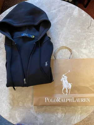 Svart hoodie från Polo Ralph Lauren - Snygg svart hoodie från Polo Ralph Lauren med dragkedja framtill och klassisk logga broderad på bröstet. Hoodien har huva med dragsko och två fickor framtill. Kan passa mindre också. Såklart äkta✅. Påsen kommer med. Om du har funderingar skriv privat 👍