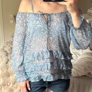 Blå paisleymönstrad blus H&M - Säljer en blå off-shoulder blus från H&M med paisleymönster och volangdetaljer. Blusen har långa ärmar med resår vid ärmslut och är gjord i ett tunt, transparent material. 