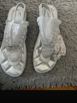 Silvriga sandaler med strassdetaljer - Snygga silvriga sandaler med glittrande strass och pärldekor framtill. De har elastiskt band runt hälen och en mjuk, vadderad sula för extra komfort. Yttersulan är mönstrad i brunt gummi. Perfekta för att lyfta din sommarstil.
