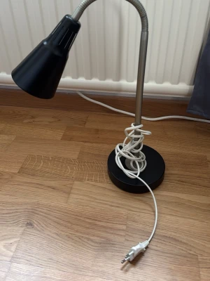 Svart skrivbordslampa med flexibel arm - Snygg svart skrivbordslampa med rund fot och flexibel silverfärgad arm som gör det enkelt att rikta ljuset. Lampan har en vit sladd med stickkontakt och en konformad lampskärm i metall. Perfekt för plugghörnan eller läshörnan.