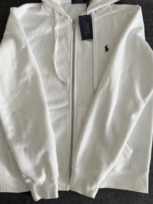 Vit hoodie från Polo Ralph Lauren - Snygg vit hoodie med dragkedja från Polo Ralph Lauren. Klassisk design med huva och dragsnören, två fickor framtill och den ikoniska lilla loggan broderad i mörkblått på bröstet. Perfekt för en stilren och avslappnad look.