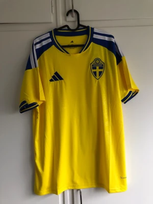 Sverige Fotbollströja Hemma 2026  - Gul svensk landslagströja från Adidas med blå och vita detaljer. Tröjan har korta ärmar, broderat landslagsemblem på bröstet och klassiska Adidas-ränder på axlarna. Tillverkad i lätt och ventilerande polyester, perfekt för fotbollsträning eller match.